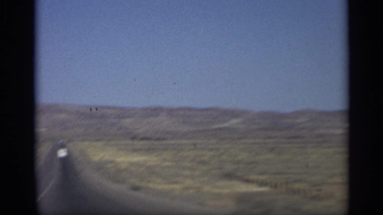 Long straight road through desert hills with a small white car in the distance.