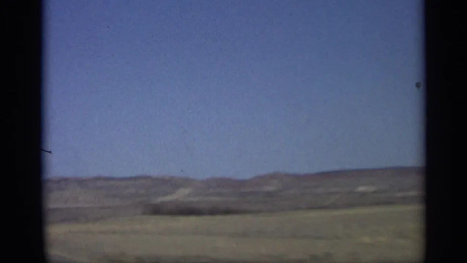 1959 Arizona home-movie frame with open arid ground, distant hills, and clear blue sky viewed from the road.