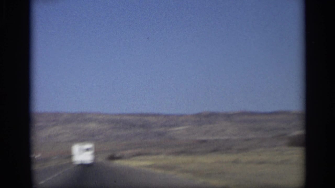 Roadway through desert hills with a distant white vehicle under blue sky.