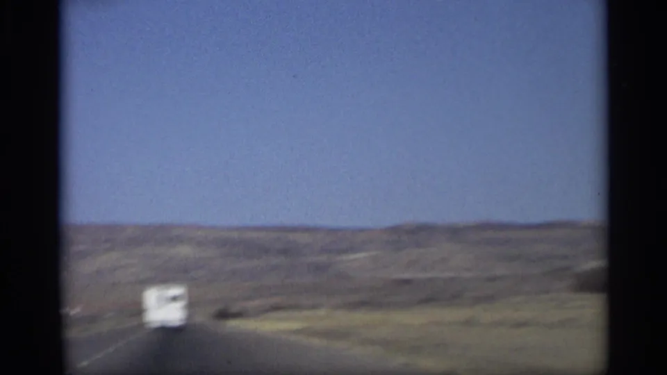 1959 Arizona 8mm frame showing a highway through low desert hills, with a small white vehicle ahead under a clear blue sky.