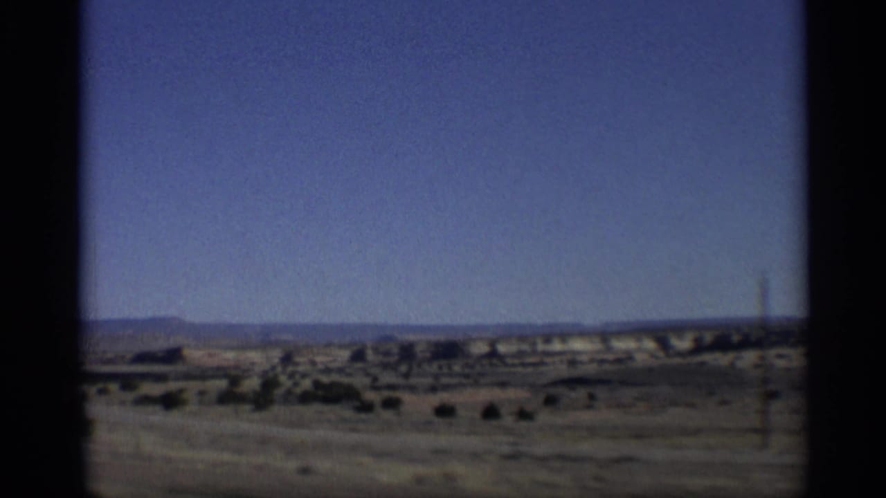 1959 Arizona home-movie frame showing arid scrubland, distant mesas, blue sky, and dark gate-edge framing from moving travel footage.