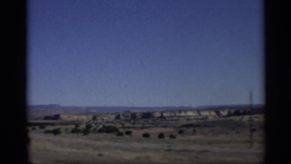 1959 Arizona home-movie frame showing arid scrubland, distant mesas, blue sky, and dark gate-edge framing from moving travel footage.