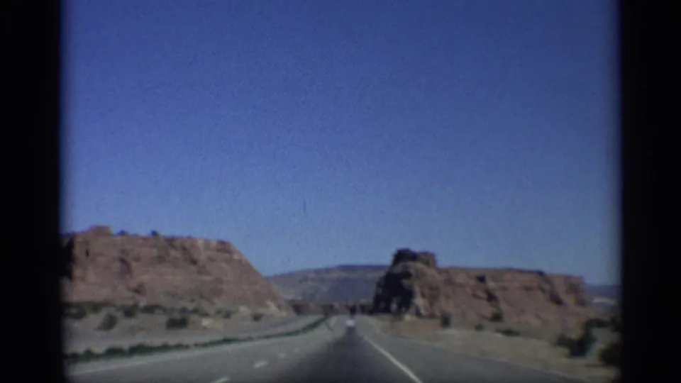 1959 Arizona frame showing a two-lane road passing between red rock formations, with sparse desert vegetation and clear sky.