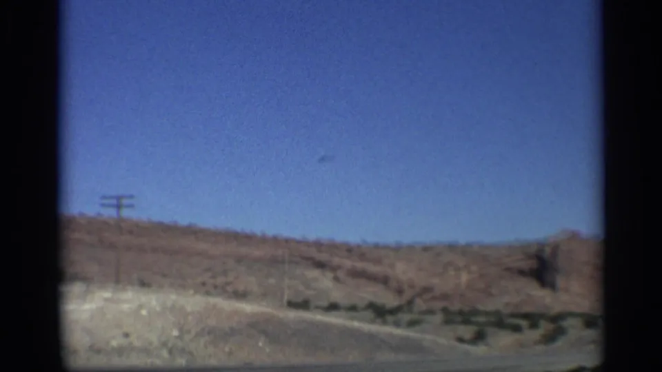 Moving 8mm view of an Arizona desert roadside, showing dry ground, low hills, utility poles, and red rock formations.