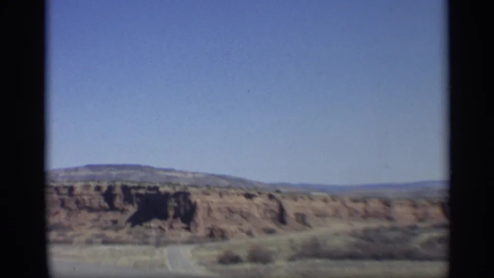 Vintage 8mm frame showing an Arizona desert road scene with reddish buttes, open pavement, scrub, and distant horizon.