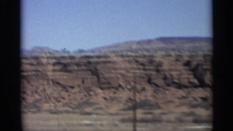 Soft, moving 8mm view of Arizona red rock terrain, with scrubby foreground and layered hills under a pale blue sky.