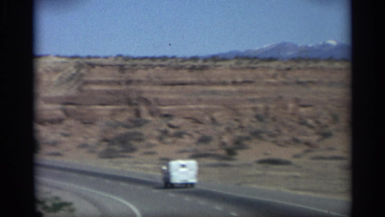 White camper vehicle on a curving desert highway with plateaus and mountains beyond.