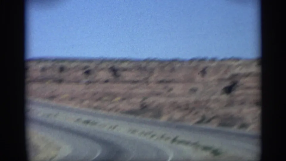 1959 Arizona 8mm frame from a moving vehicle, showing a curving road and soft red rock desert slope.