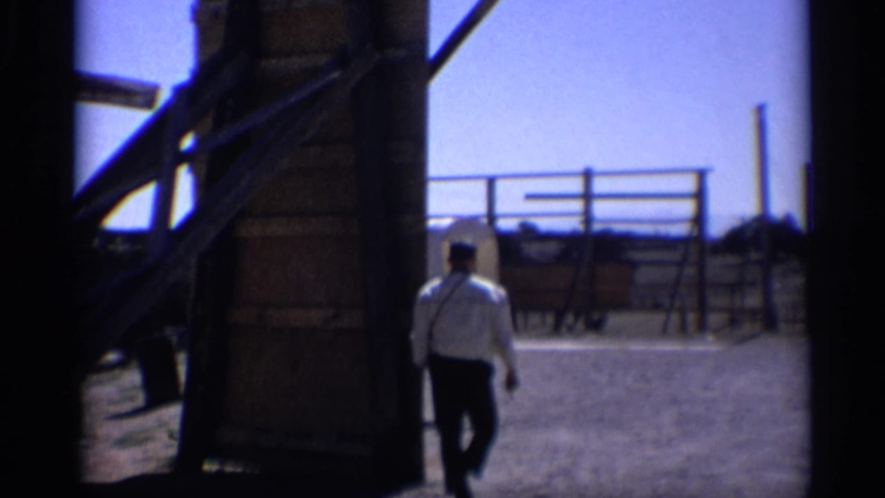In 1959 Arizona 8mm footage, a man walks out from a shadowed structure toward fencing and a western display area.