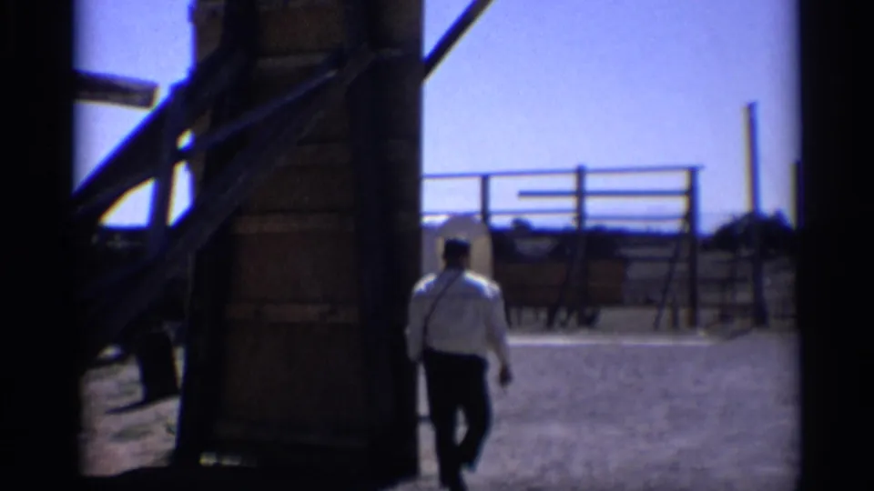 In 1959 Arizona 8mm footage, a man walks out from a shadowed structure toward fencing and a western display area.