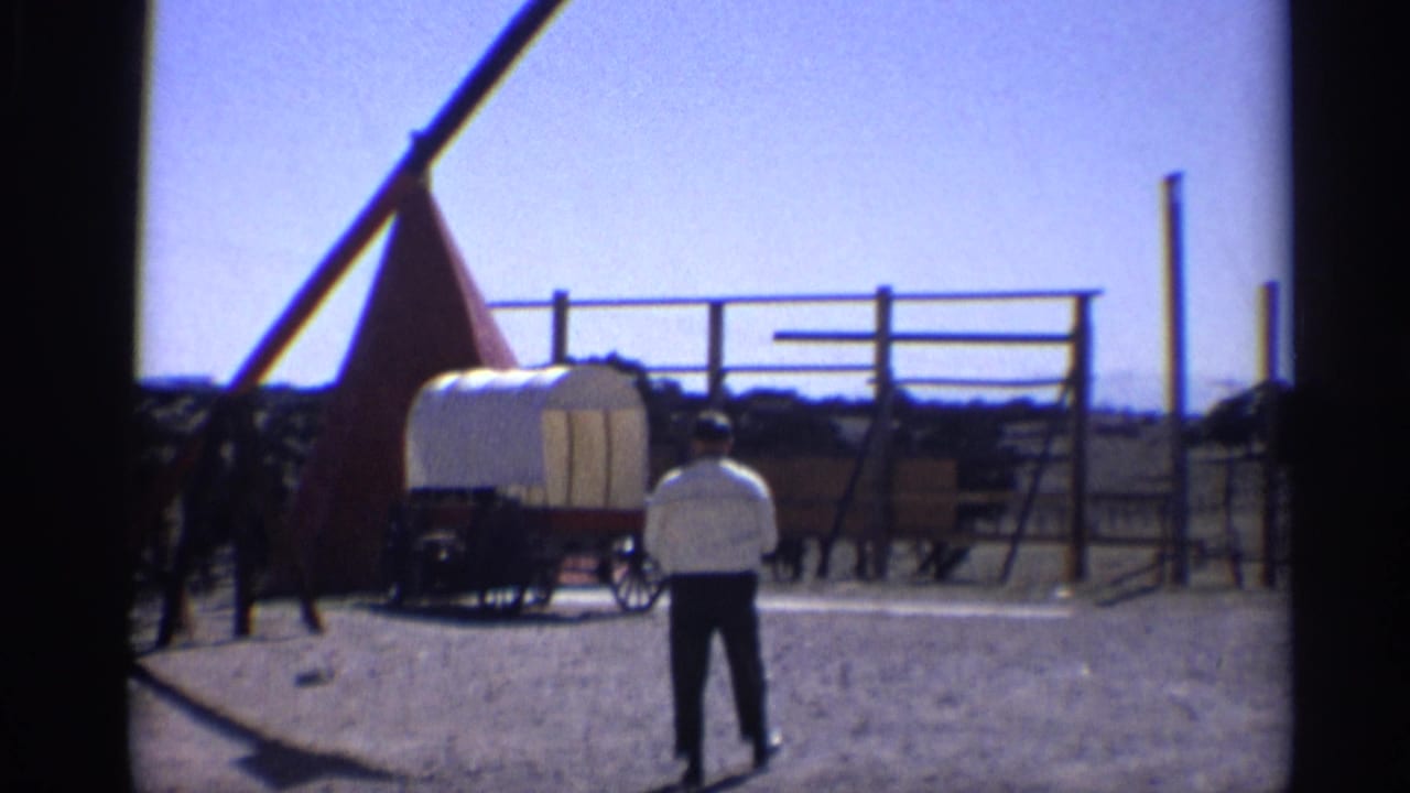 Man stands near a covered wagon and wooden fence in bright sunlight.