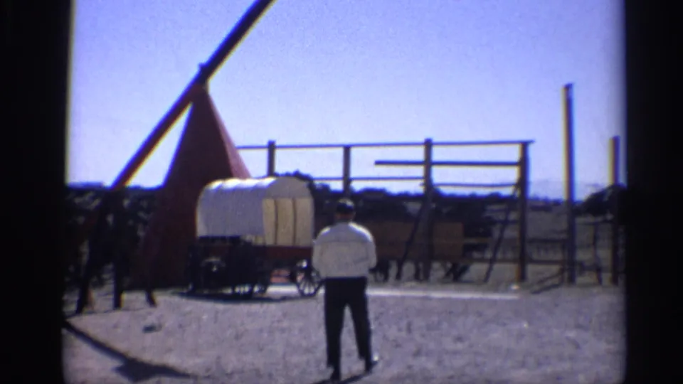 A man in a white shirt stands near a covered wagon, fence rails, and dark animals in a sunlit Arizona historic-site setting.