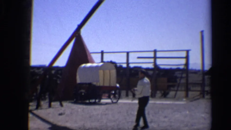 The man passes the covered wagon in a bright 1959 Arizona 8mm frame with fencing, posts, and a western-themed display area.