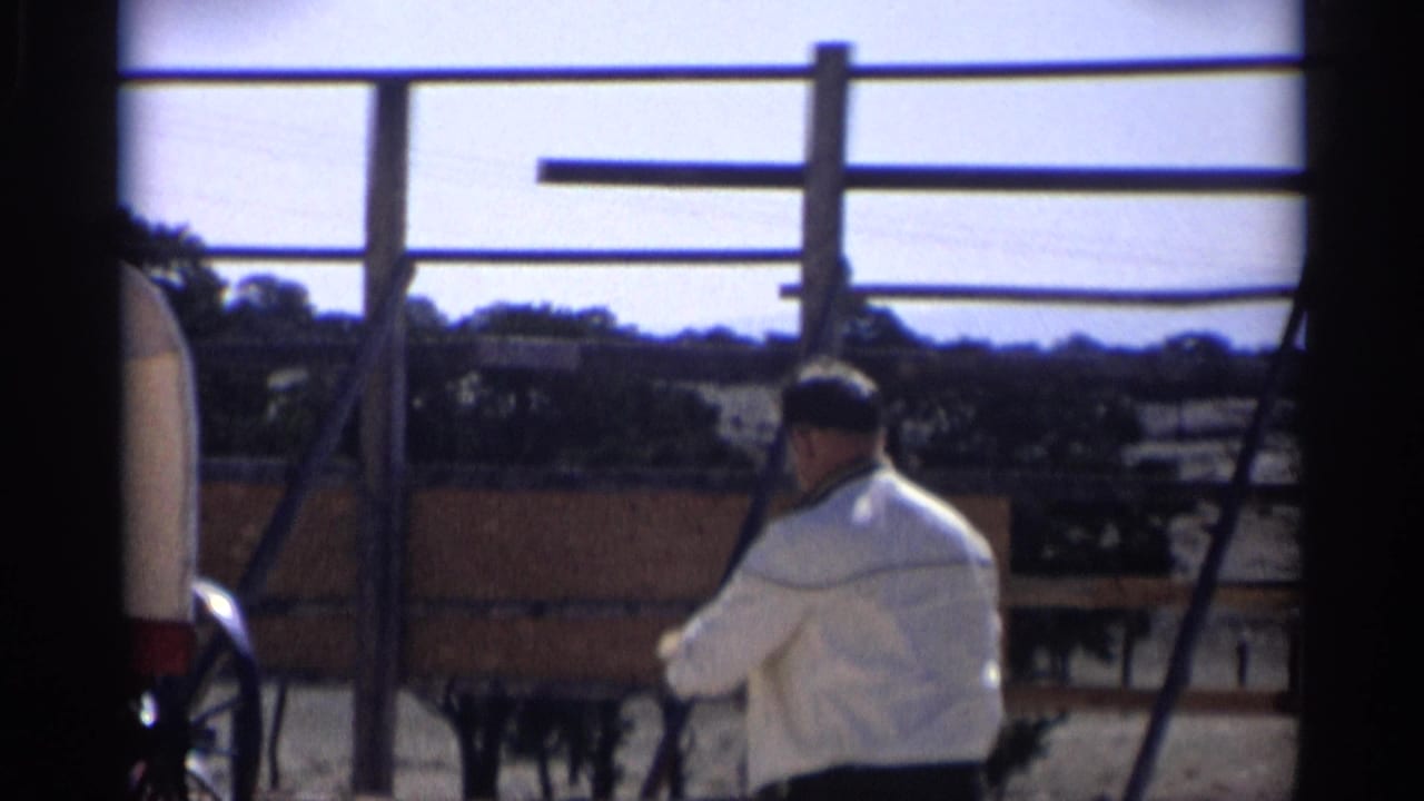 Adult man in a light jacket stands beside fencing with a covered wagon partly visible.