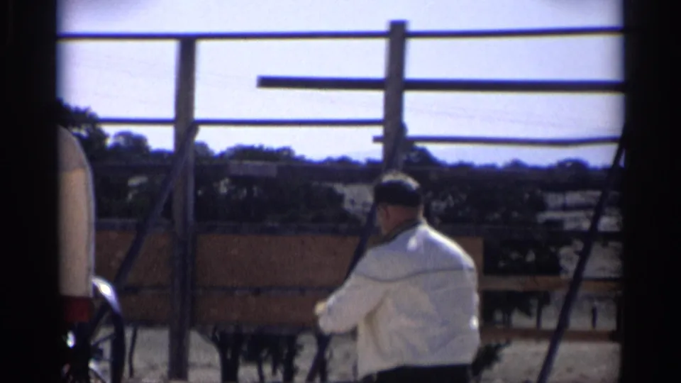1959 Arizona ranch scene with an adult man in a light jacket near a covered wagon, fence rails, and distant terrain.