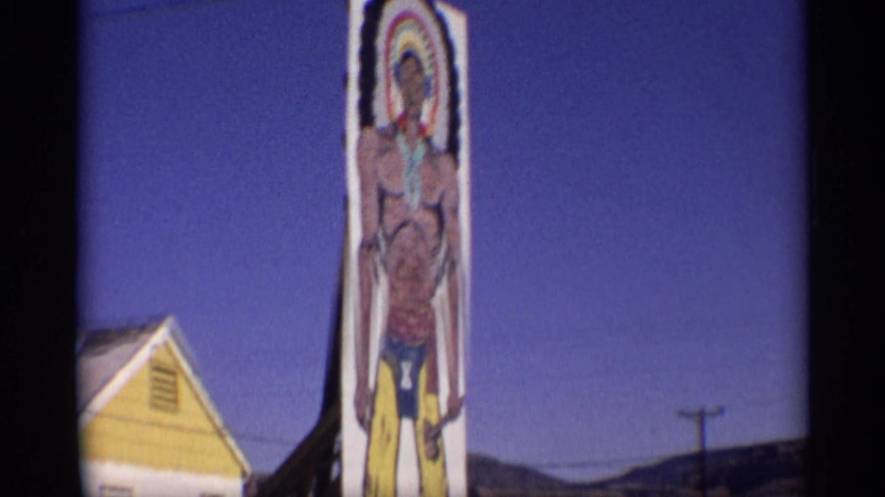 1959 Arizona 8mm frame showing a tall painted Native American figure sign next to a yellow and white roadside gift shop building.