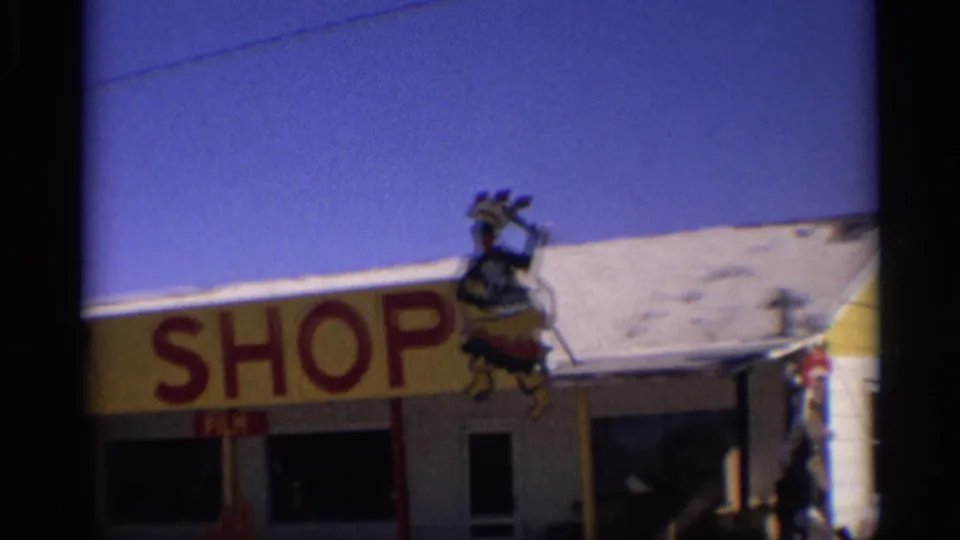 1959 Arizona 8mm frame showing the gift shop facade with large red SHOP lettering, roof-mounted figure, windows, and blue sky.
