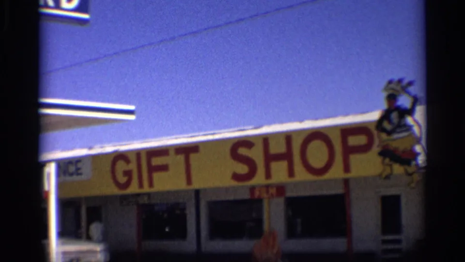1959 Arizona frame of a roadside storefront with large GIFT SHOP lettering, windows, yellow facade, and roof-mounted figure.