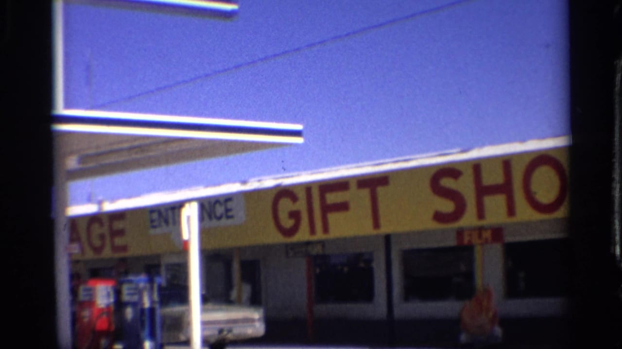 Gas station storefront with yellow gift shop sign, pumps, canopy, and parked car.
