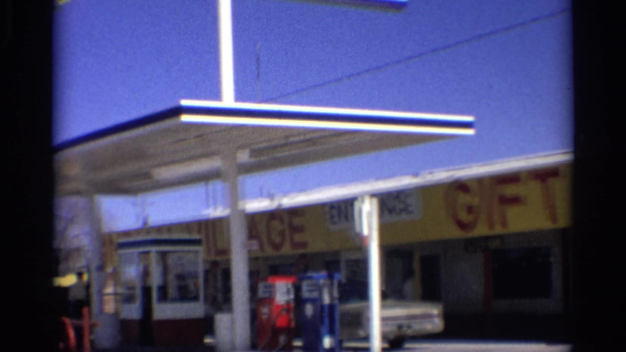 1959 Arizona roadside station view with canopy supports, gift shop lettering, pumps, entrance, and a white car near the storefront.