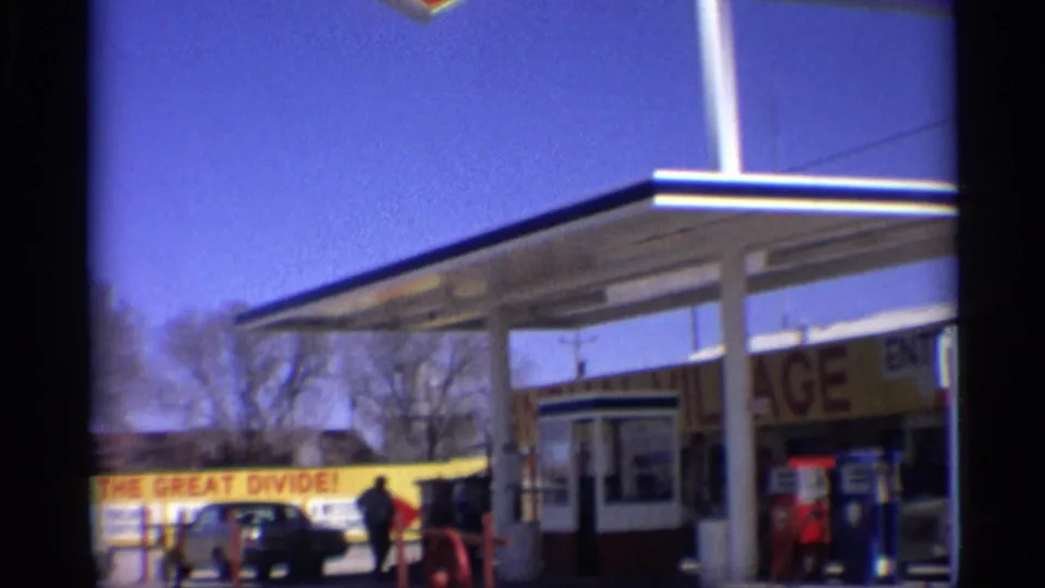 1959 Arizona frame showing the station forecourt, Welcome to the Great Divide sign, dark car, walking person, pumps, and bare trees.