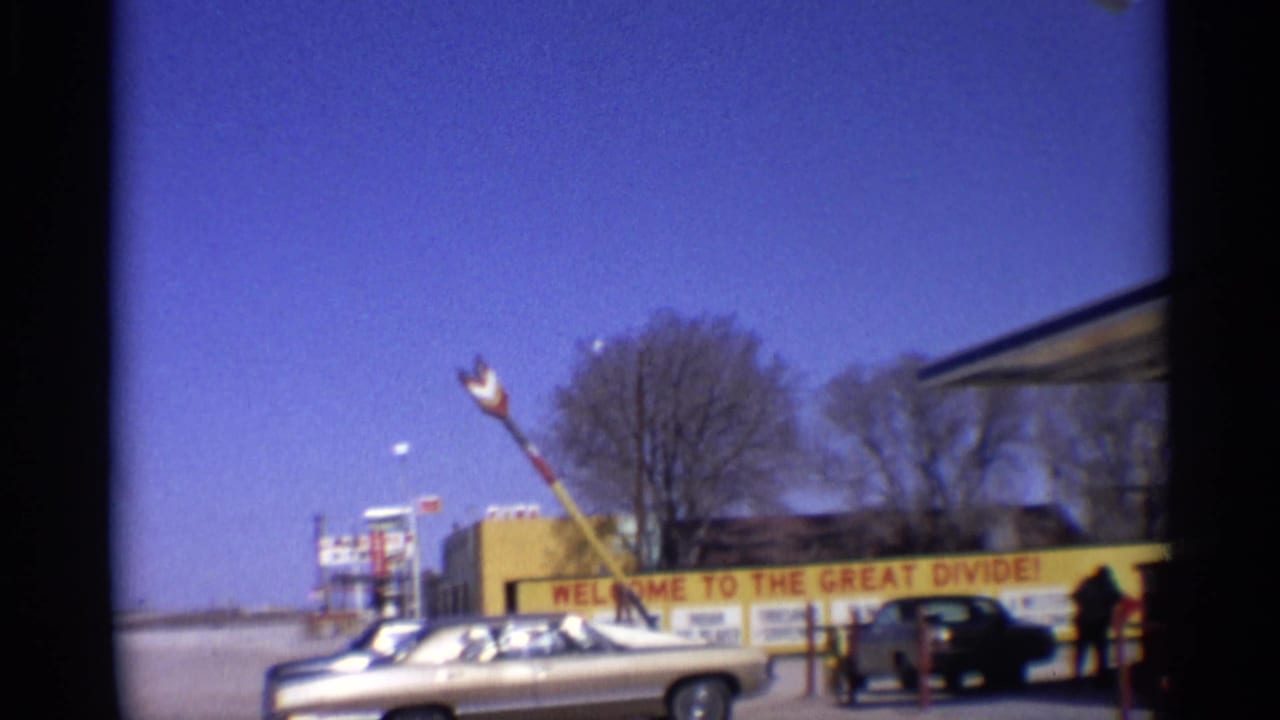 1959 Arizona road-trip frame showing a gold convertible beside a roadside welcome sign, with travelers and a large arrow sculpture nearby.