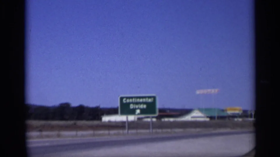 1959 Arizona frame centered on a green Continental Divide road sign, with highway lanes and a service station beyond it.