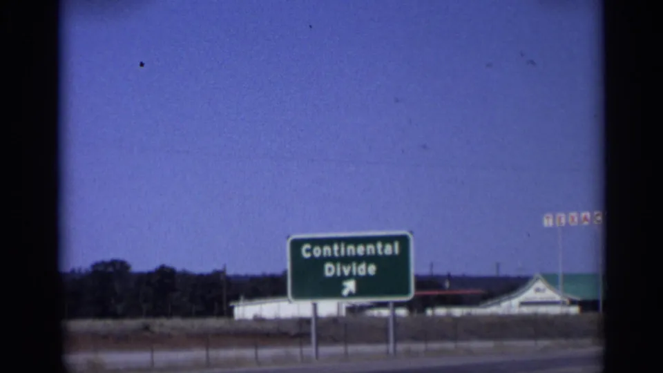 1959 Arizona roadside view of a Continental Divide sign with an arrow, highway pavement, and low service-station buildings beyond.