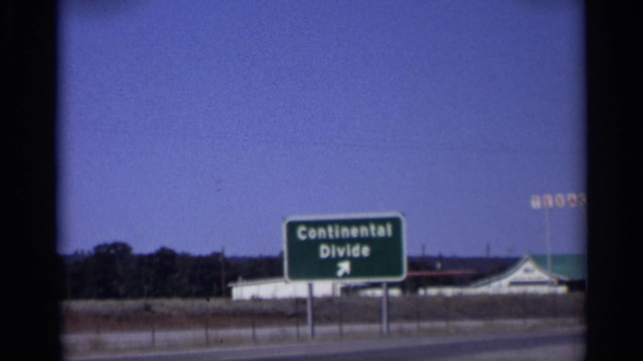1959 Arizona home-movie frame showing a green Continental Divide directional sign with an arrow near a roadside building and distant service signage.