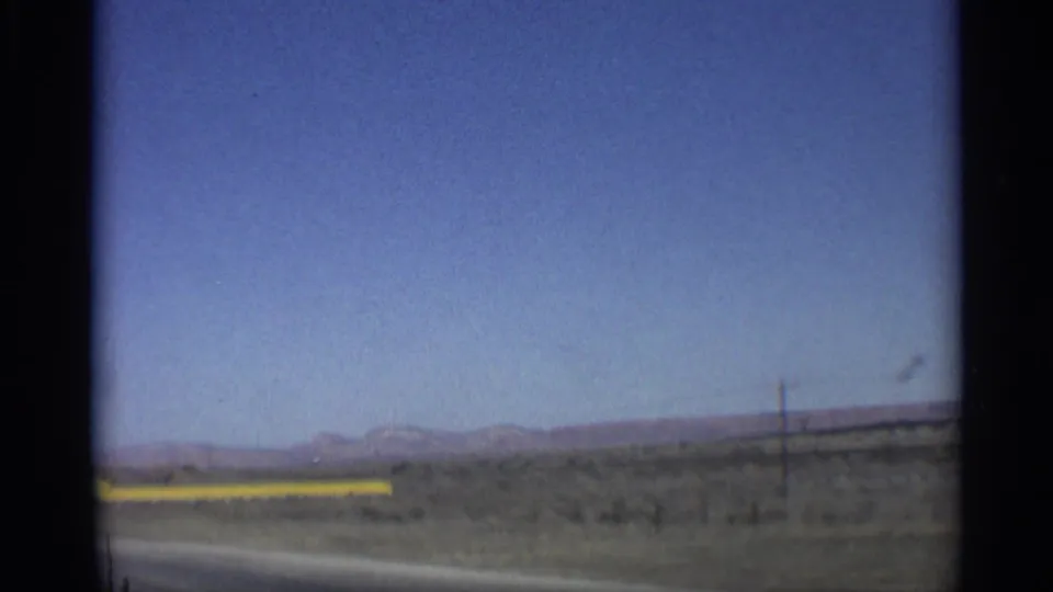 1959 Arizona roadside landscape showing a yellow guardrail, open desert ground, utility poles, and low mountains beneath a wide blue sky.