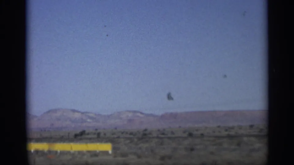 Final 1959 Arizona road-trip frame looking across scrub desert toward low mountains, with a yellow roadside barrier and a small bird-like shape in the sky.