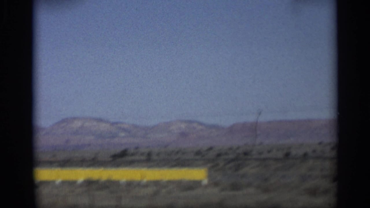 1959 Arizona 8mm frame showing a yellow roadside barrier across desert scrub with low mountain ridges in the distance.