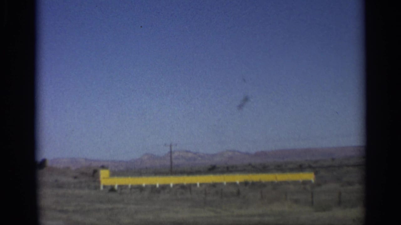 Yellow roadside barrier, utility pole, desert scrub, and mountains beneath a clear sky.