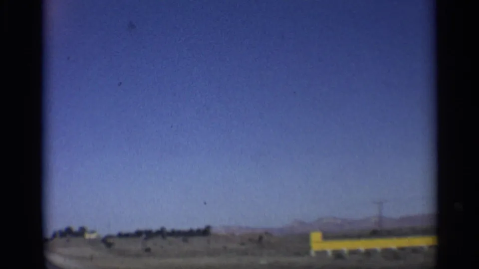 1959 Arizona home-movie frame looking across an open roadside toward a yellow barrier, power pole, desert plain, and mountains.