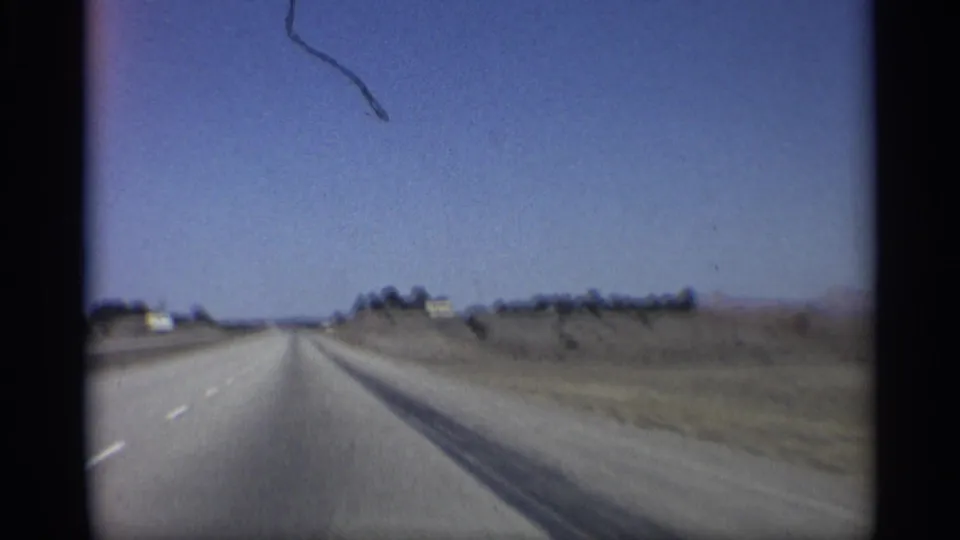 1959 Arizona 8mm frame showing a moving-vehicle view down an open highway with sparse roadside development and distant mountains.