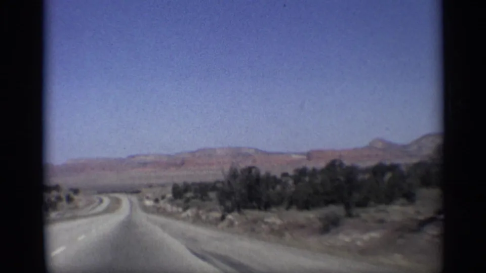 Arizona 8mm travel frame showing a highway bend, sparse roadside vegetation, distant hills, and small vehicles ahead.