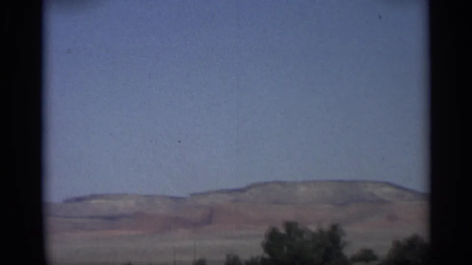 1959 Arizona 8mm frame showing a broad desert plain, low hills, scattered shrubs, and a clear blue sky.