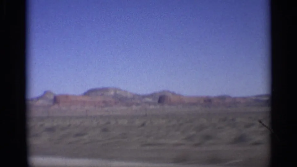1959 Arizona 8mm frame showing open desert, scrub vegetation, and distant red rock formations from a moving vehicle.