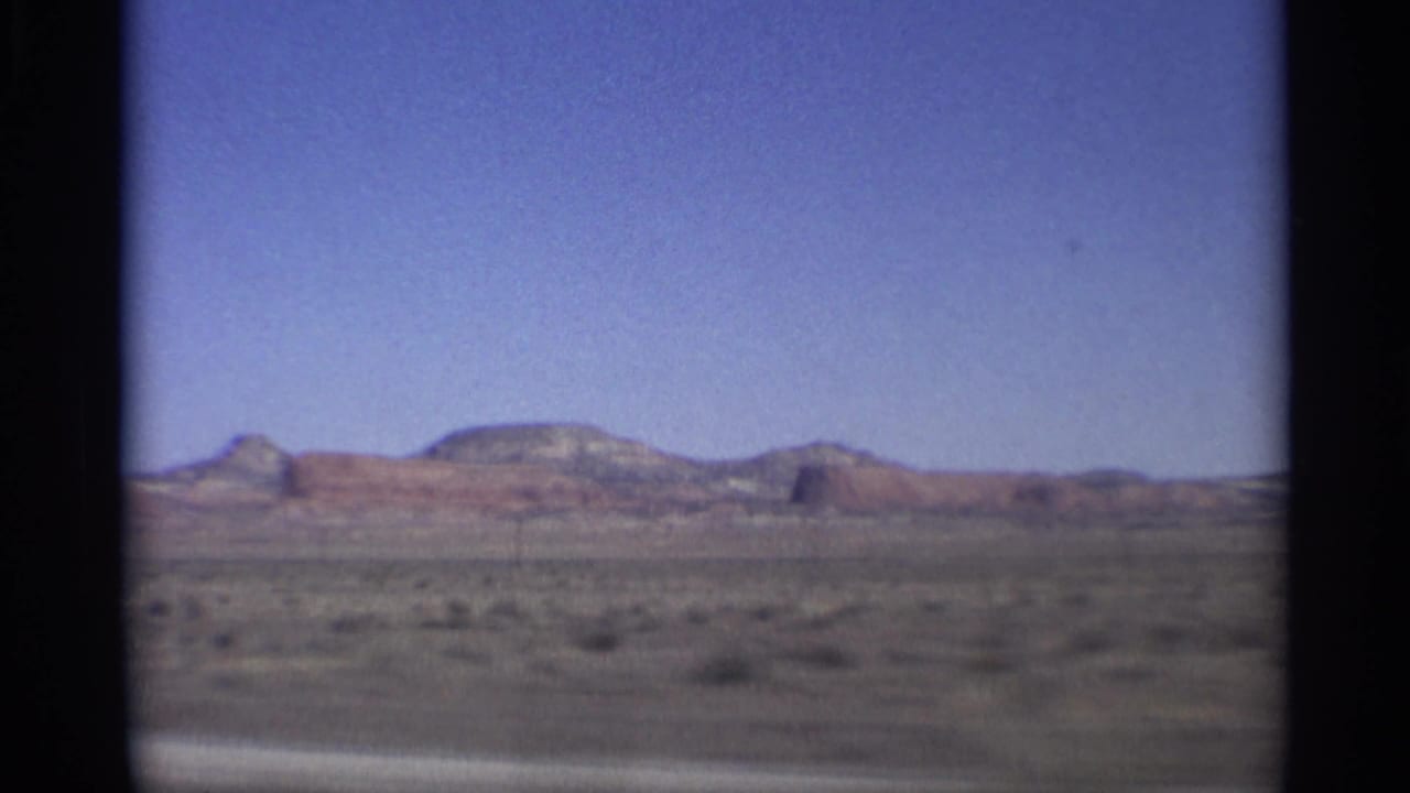Desert plain with distant red rock mesas under a blue sky, framed by 8mm film gate edges.