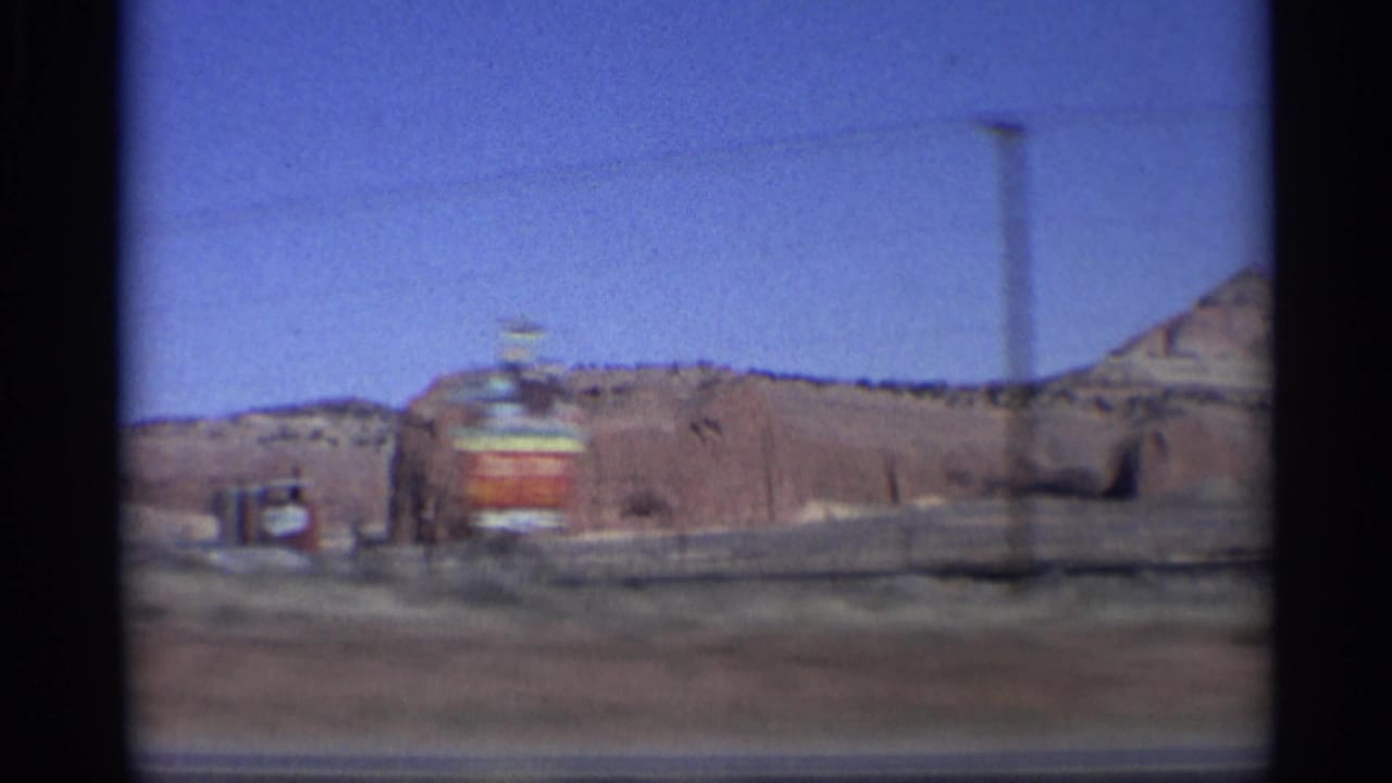 1959 Arizona desert roadside view with red rock terrain, a small distant structure, and a utility pole seen through 8mm film blur.