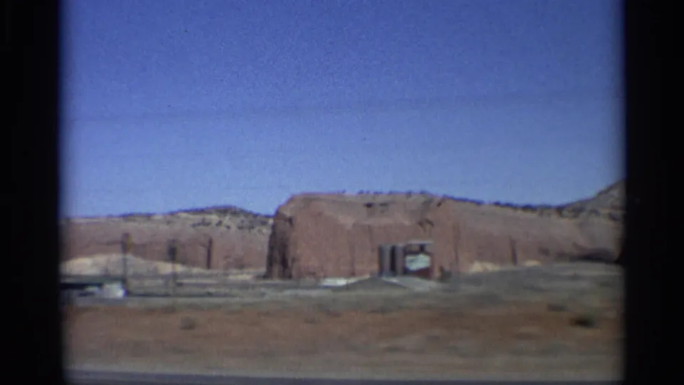 1959 Arizona 8mm frame showing a low red rock formation, open desert ground, road edge, and clear sky from a passenger viewpoint.