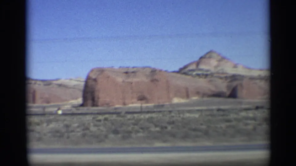 1959 Arizona desert landscape with a red rock butte, roadside barrier, scrubland, and clear sky captured on color 8mm film.