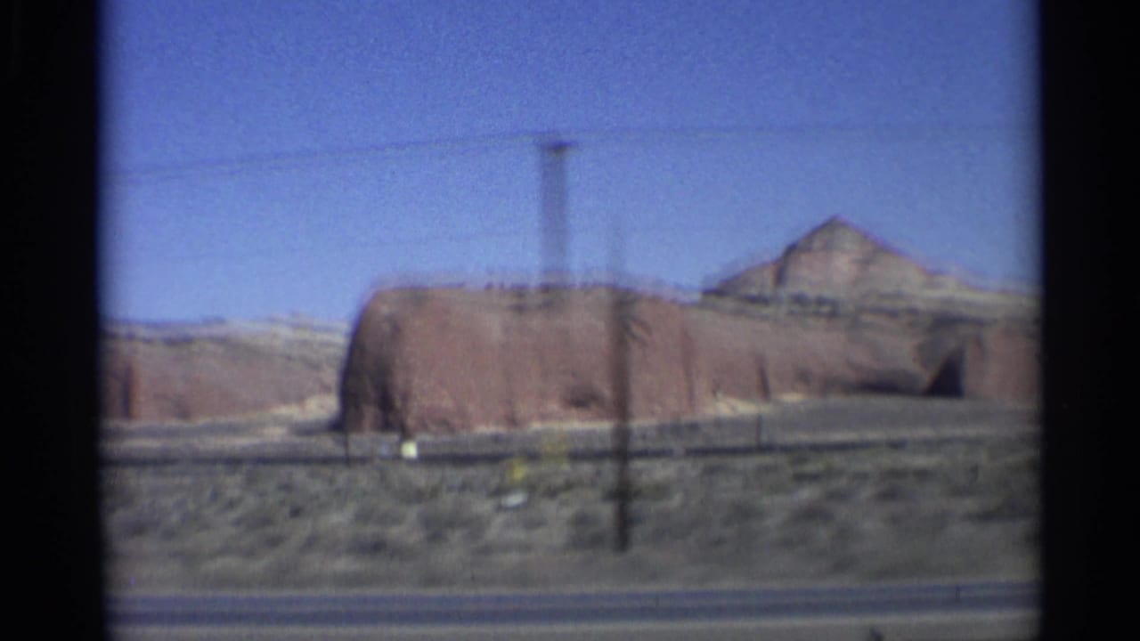 1959 8mm Arizona road-trip frame showing a red sandstone formation beyond a roadside shoulder, with desert scrub, a utility pole, and blue sky visible from a moving vehicle.