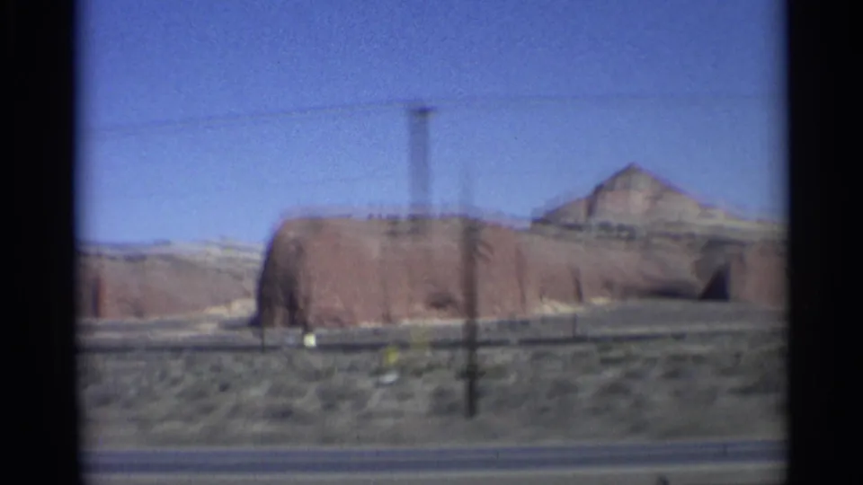 1959 8mm Arizona road-trip frame showing a red sandstone formation beyond a roadside shoulder, with desert scrub, a utility pole, and blue sky visible from a moving vehicle.