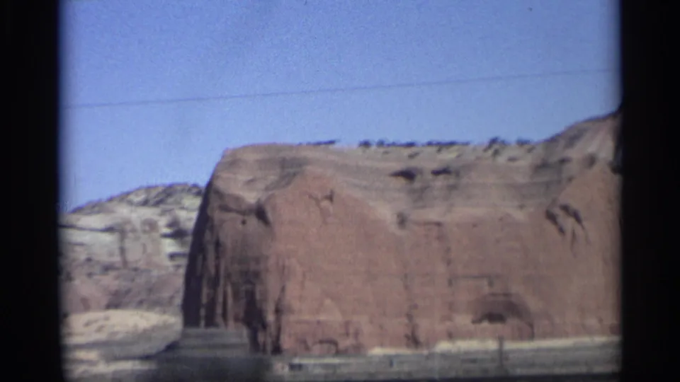 1959 8mm Arizona road-trip frame showing a closer view of a broad red sandstone wall, pale desert strata, sparse roadside ground, and a clear blue sky.