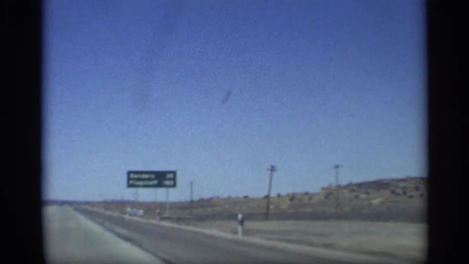 1959 8mm Arizona highway frame showing an open road, utility poles, desert terrain, and a green distance sign listing Sanders and Flagstaff beneath a clear blue sky.