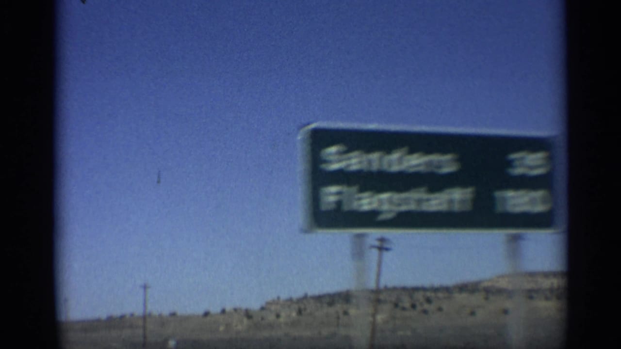 A close, motion-blurred 1959 Arizona highway sign for Sanders and Flagstaff passes during a moving-car shot.