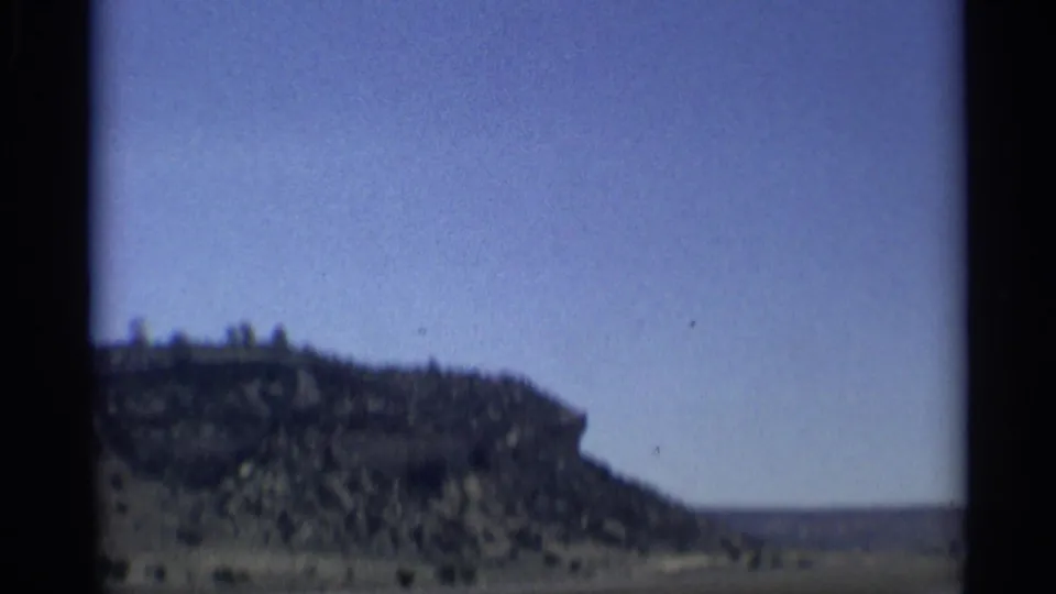 A soft 8mm view of Arizona desert terrain, with a rocky slope, sparse brush, and broad blue sky from a moving vehicle.