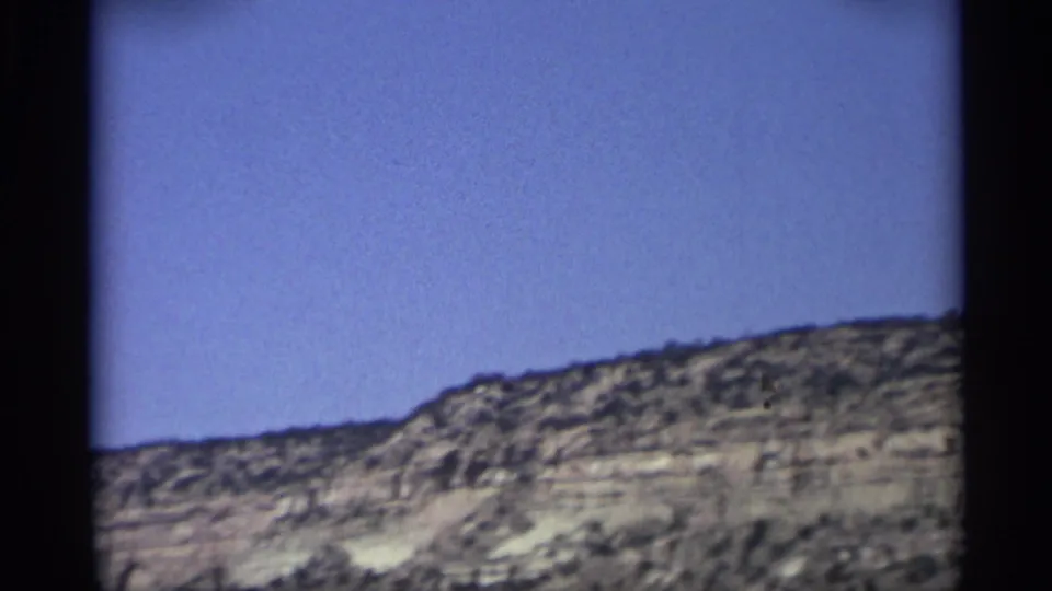 1959 Arizona 8mm frame showing a low rocky ridge and open blue sky from a moving roadside viewpoint.
