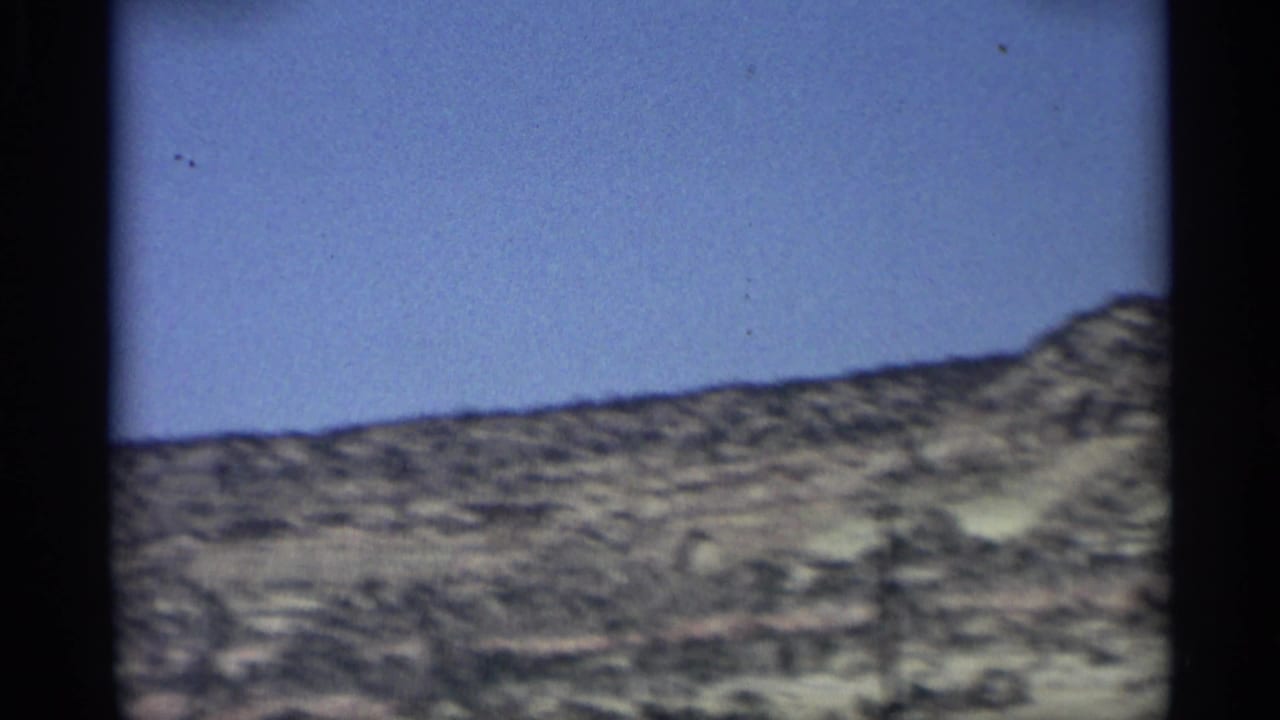 Dry desert hillside under a blue sky, filmed from a moving vehicle.